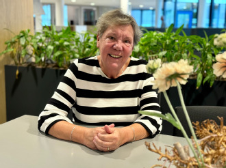 Til van den Berg. Ze draagt een zwart-wit gestreepte trui. Ze zit aan een tafel en er staat een grote bloem op de tafel.